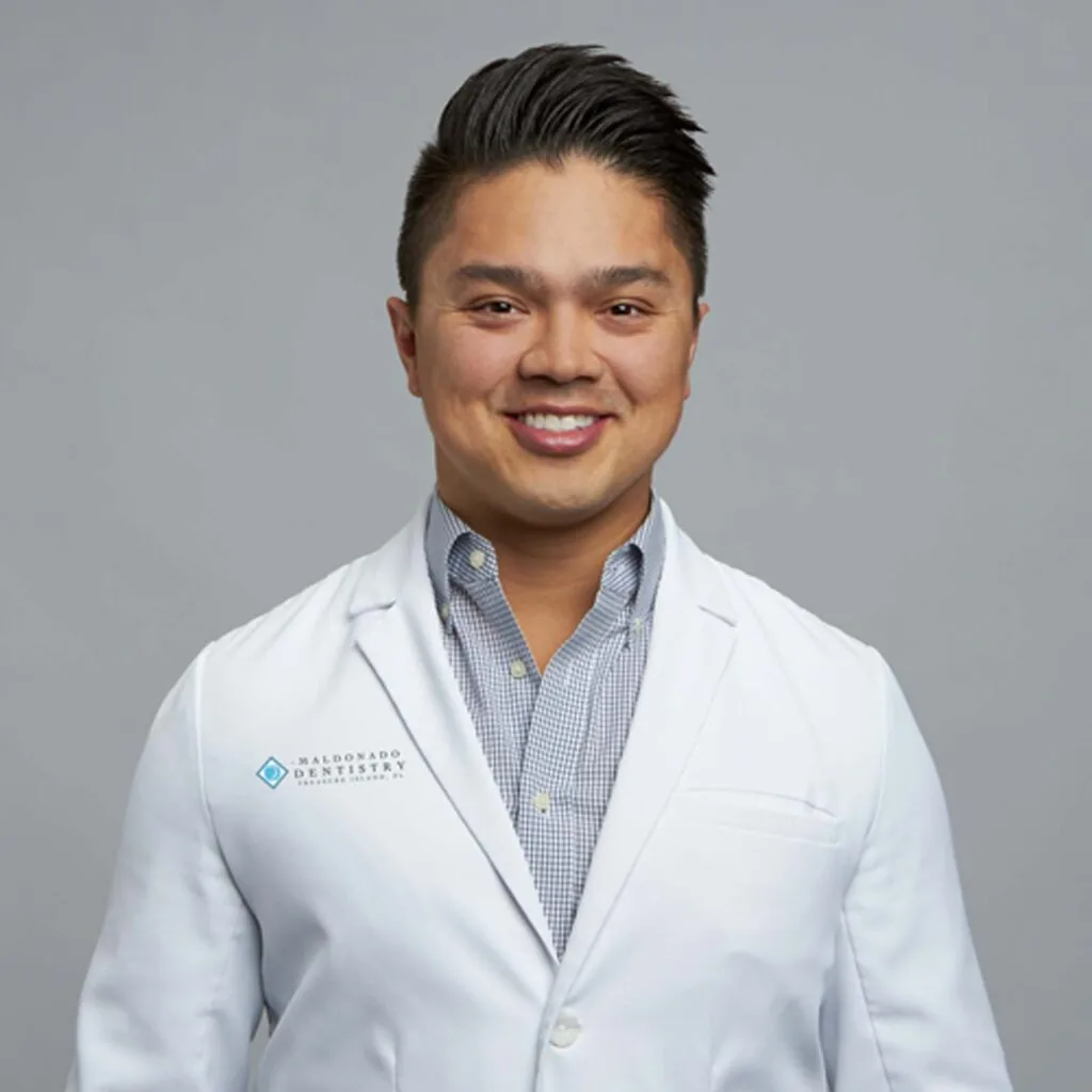 Dr. Alex Maldonado wearing a white lab coat with a 'MALDONADO DENTISTRY' logo and a small blue diamond-shaped icon on the left side, over a light blue checkered shirt. The background is plain white.