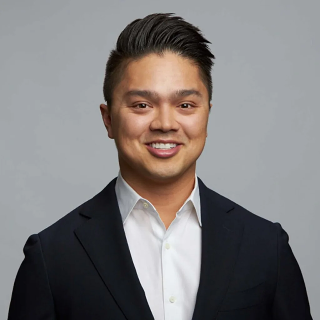 The image shows Dr. Alex Maldonado wearing a dark suit jacket over a white dress shirt. The background is plain and gray.